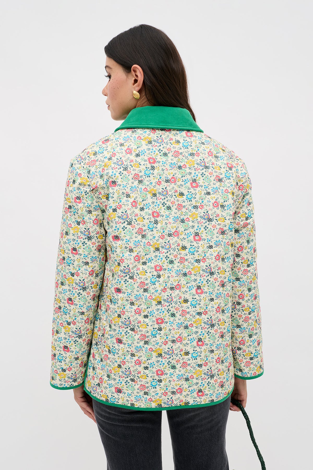A woman with long dark hair and yellow earrings faces away, wearing the Earla - Amalfi Floral Cream jacket with a bright green collar and cuffs, paired with dark pants, set against a plain light background.
