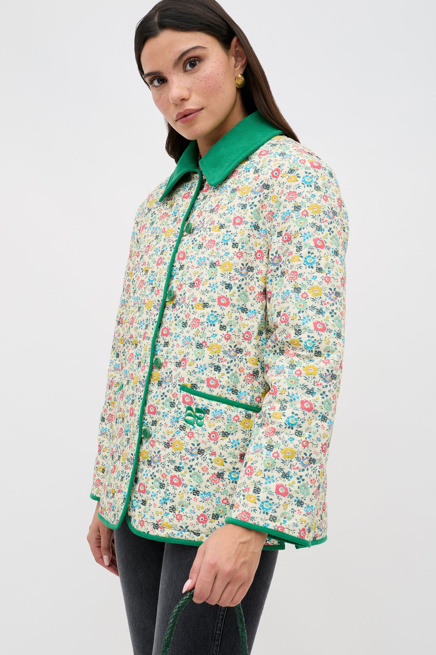 A woman wearing the Earla - Amalfi Floral Cream quilted jacket with green trim stands against a plain white background, paired with dark jeans and holding a green purse.