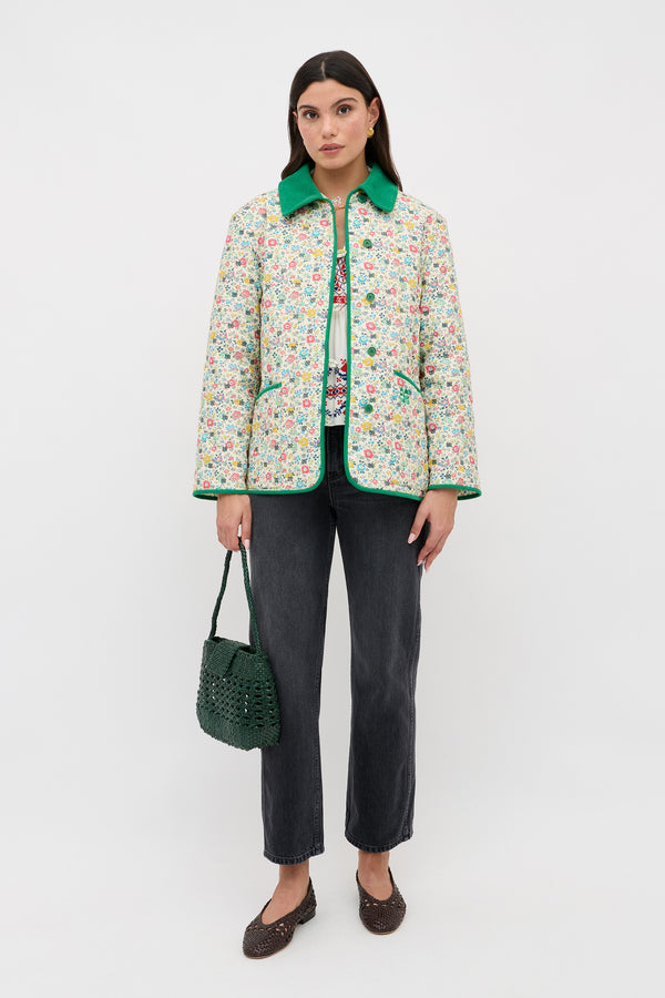 A woman with straight dark hair and a neutral expression stands against a white background wearing the Earla - Amalfi Floral Cream jacket, dark jeans, brown woven shoes, and holding a green woven handbag.