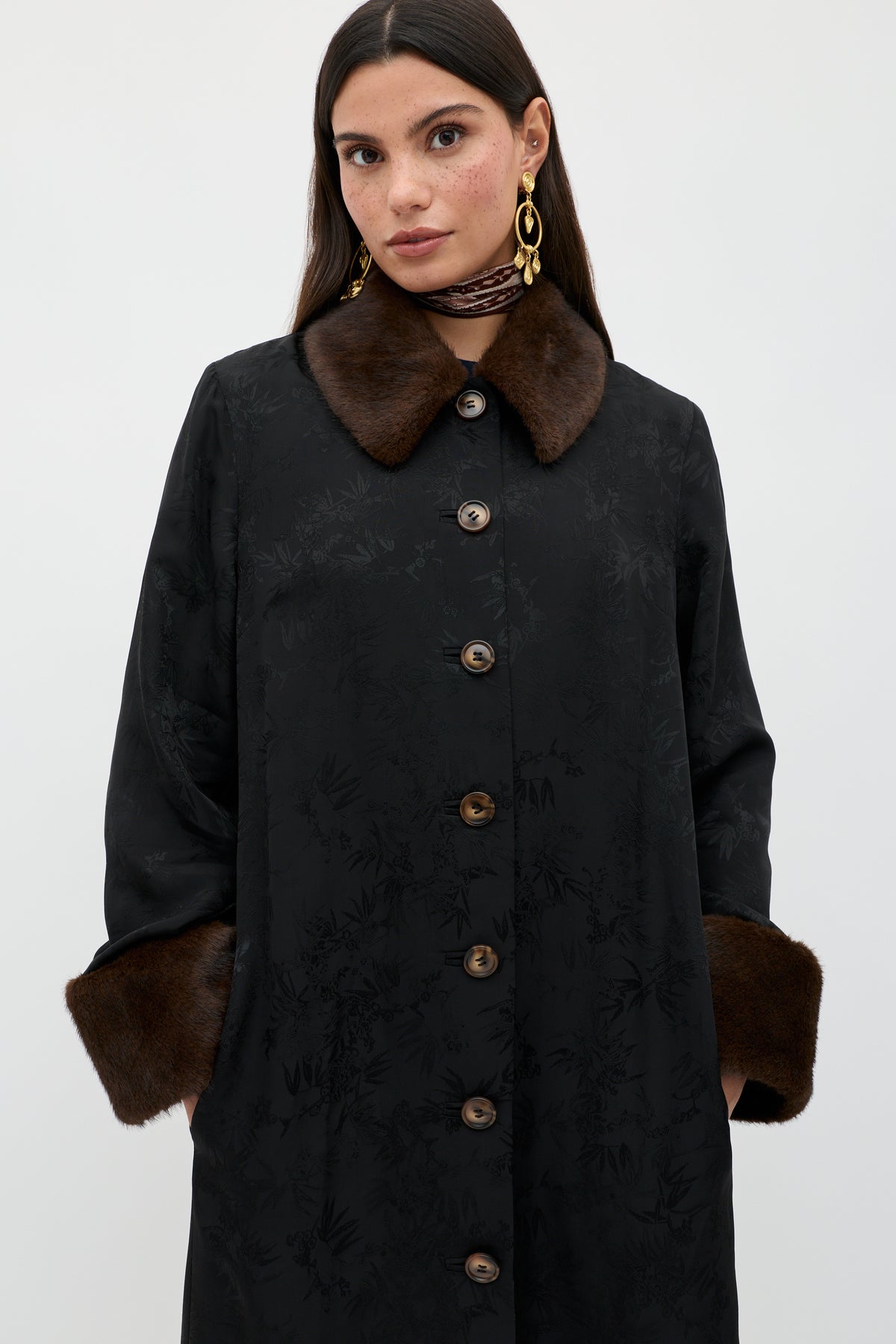 A woman wearing the Jonah - Black coat with brown fur collar and cuffs, gold earrings, and a patterned neck scarf stands against a plain light background.