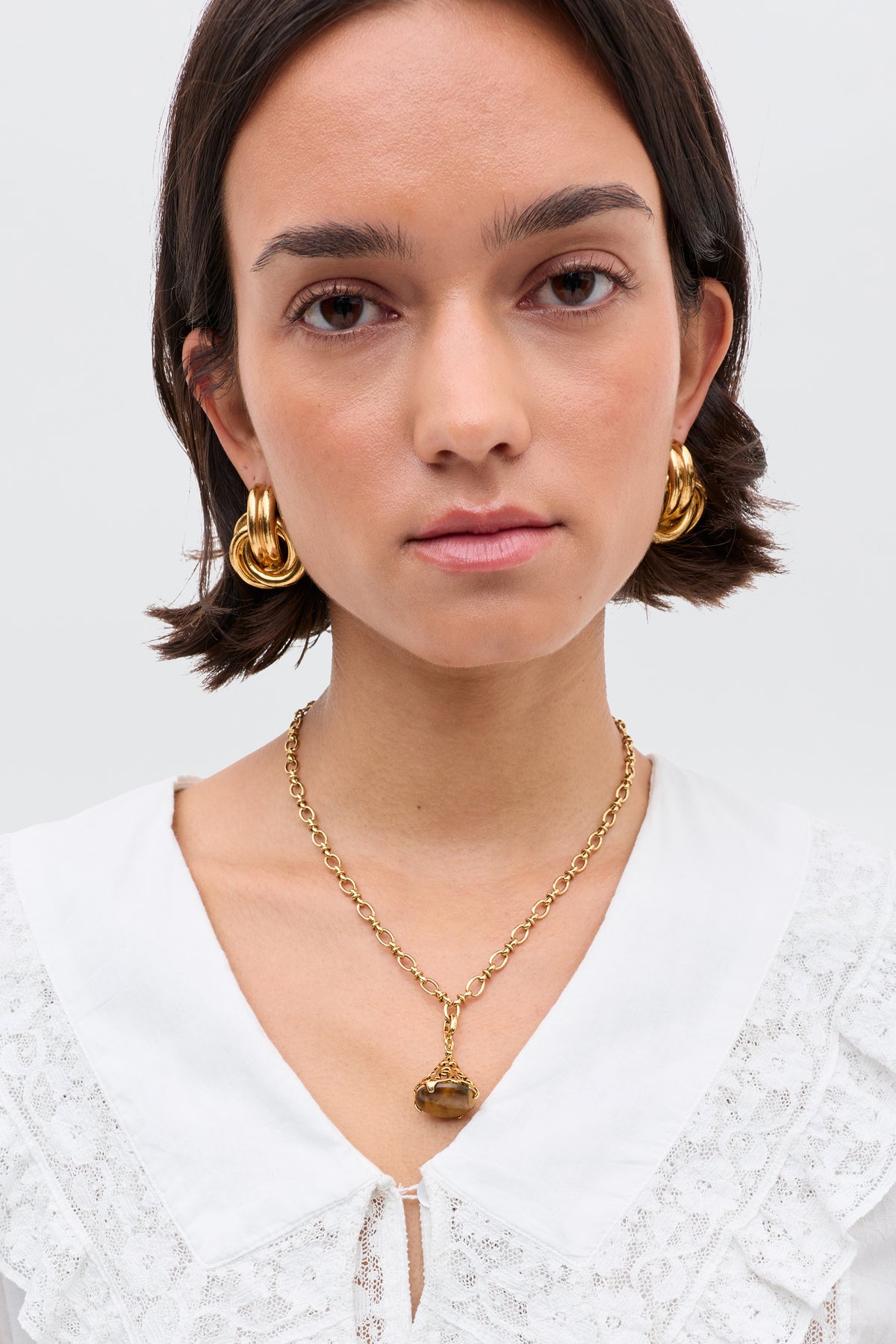 A woman with short dark hair wears Maella - Gold chunky 18-karat gold-plated hoop earrings, a matching filigree charm necklace, and a white lace-detailed blouse, standing against a plain light background.