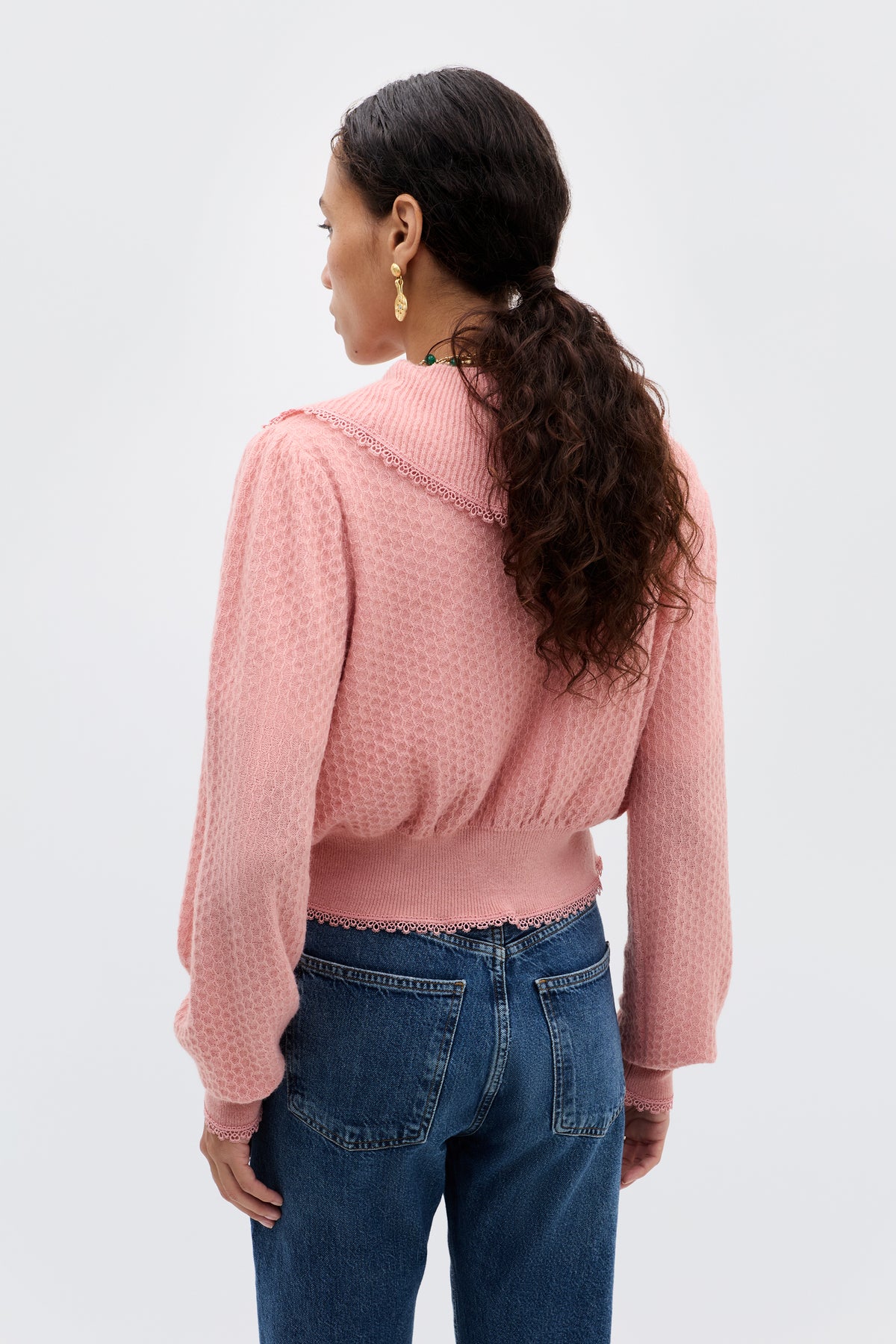 A woman with long, curly hair in a ponytail wears the Kianna - Blush textured pink transitional knit sweater with lace trim and blue jeans, standing with her back to the camera against a plain light background.