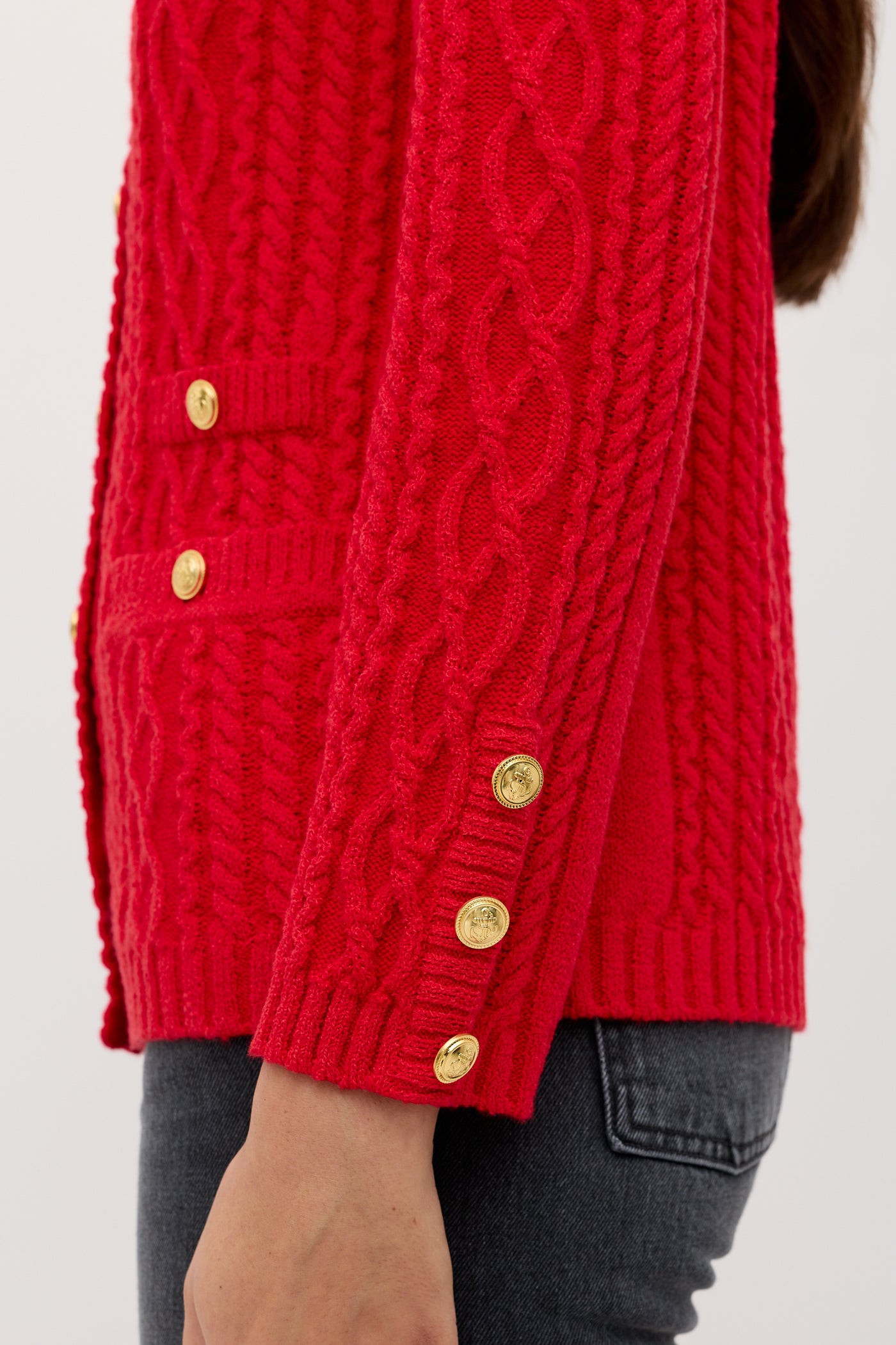 A person is seen from the side wearing the “Maurya - Red” textured cable-knit sweater with gold buttons on the sleeve and pocket, paired with dark gray jeans. The photo highlights the sweater’s detailed design.
