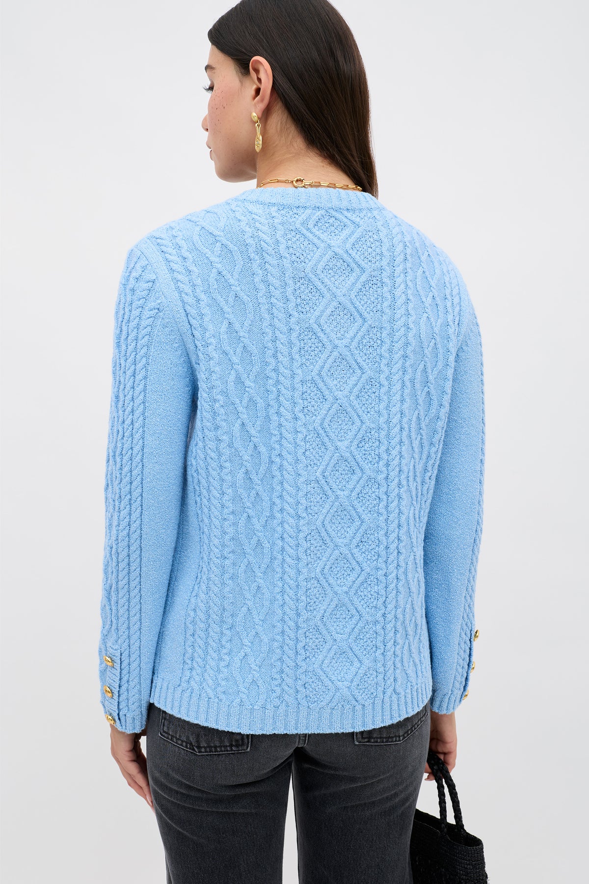 A person with long dark hair, gold earrings, and dark jeans is shown from the back against a plain light background, wearing the Maurya - Dusty Blue cable-knit sweater with diamond and braid patterns.