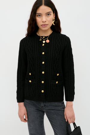 A woman with long dark hair stands against a white background wearing the Maurya - Black cable-knit cardigan with gold buttons, dark jeans, layered gold necklaces, and holding a black textured bag.