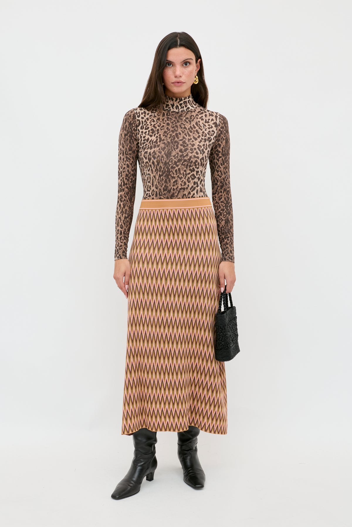 A woman stands against a white background wearing the Polo - Leopard Brown turtleneck, a long zigzag-patterned skirt, black knee-high boots, and holding a small black handbag.