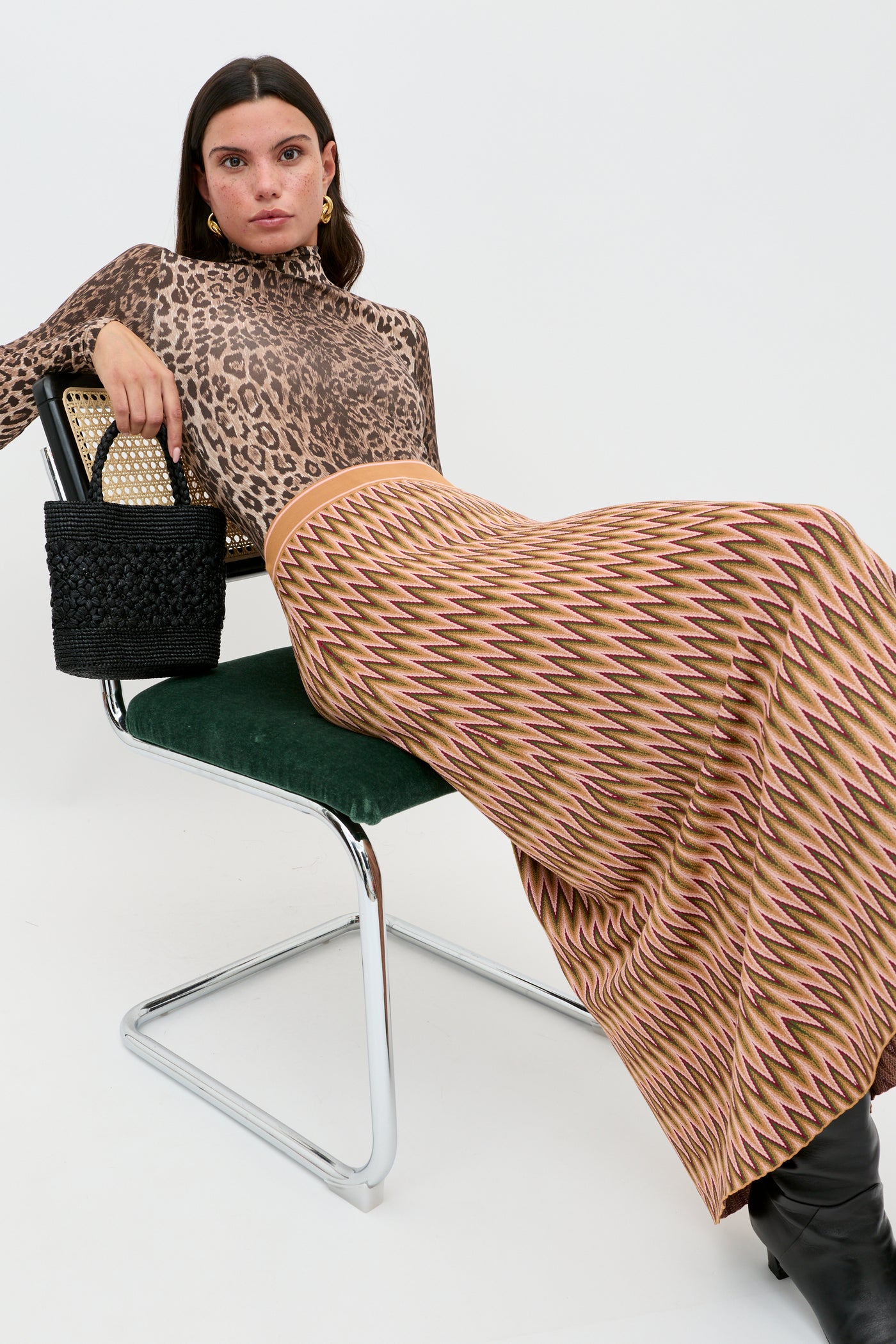 A woman sits on a modern chair in the Polo - Leopard Brown TENCEL Modal turtleneck, a long zigzag skirt, black boots, and a woven black handbag, looking directly at the camera against a plain white background.