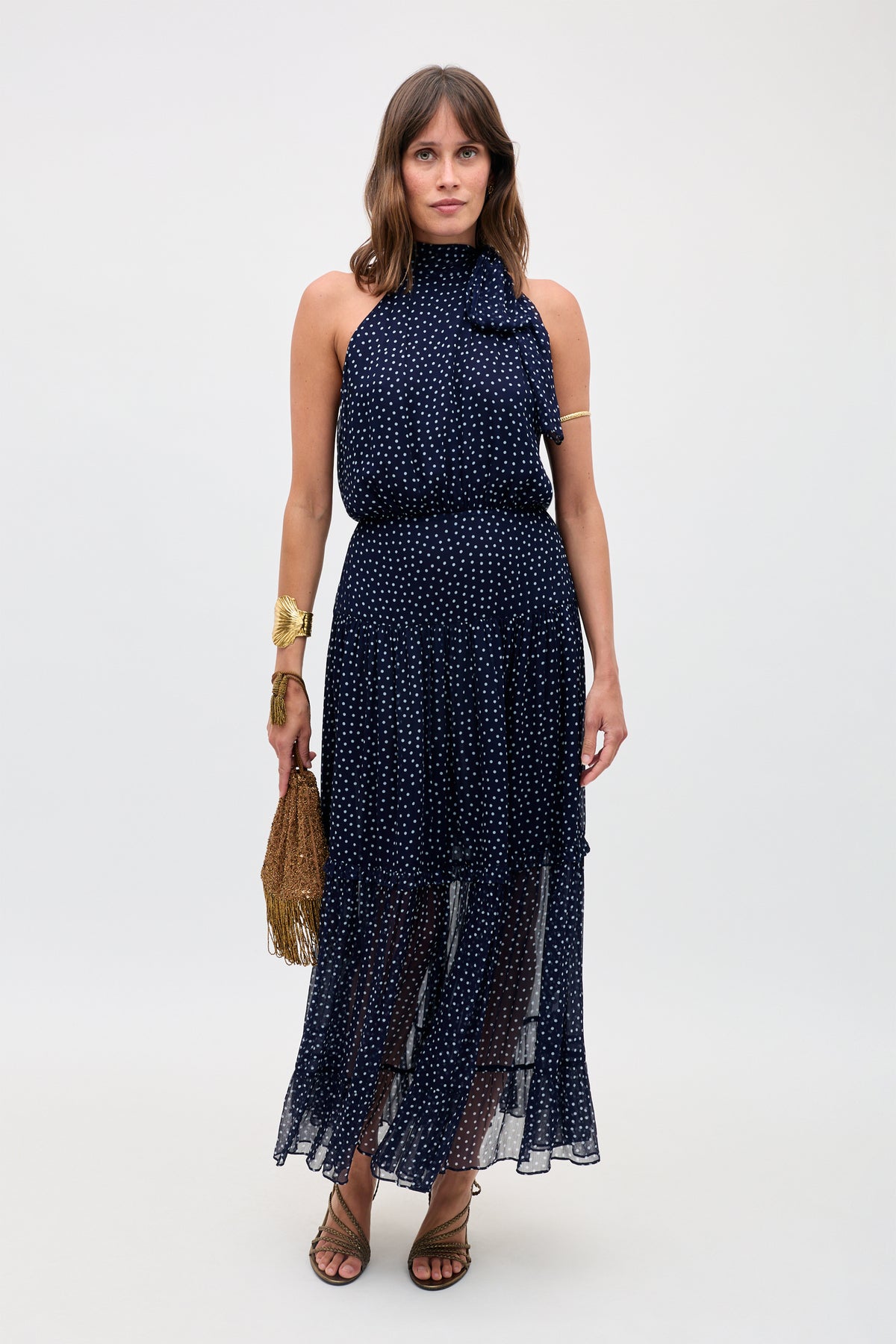 A woman stands against a plain background, wearing the Abi - Mini Navy Polka Dot dress with white polka dots. She accessorizes with a gold fringe handbag, gold jewelry, and brown strappy sandals.