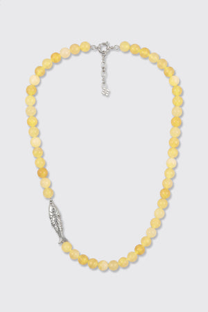 The Amoura - Lemon necklace is made of round yellow beads and features a silver fish-shaped charm on one side, along with a silver clasp and an adjustable chain.