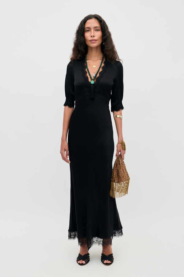 A woman wearing the Annina - Black midi dress with lace trims, cut on the bias for a flattering silhouette, paired with black sandals, layered necklaces, a gold bracelet, and holding a gold fringe handbag, stands against a plain white background.