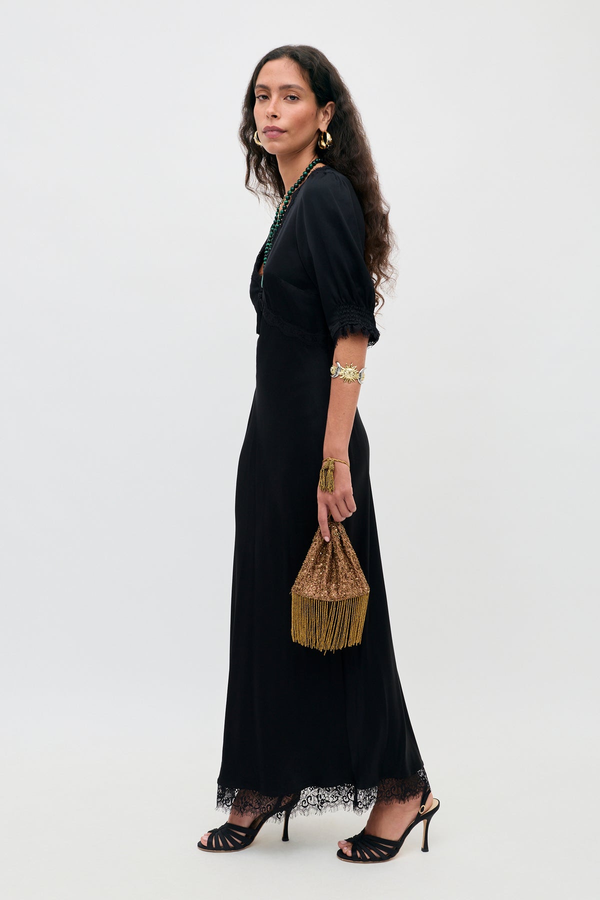 A woman in the “Annina - Black” midi dress with delicate lace trims, paired with black heels and gold jewelry, holds a gold fringed bag. She stands sideways against a plain white background, her long wavy hair framing a composed expression.