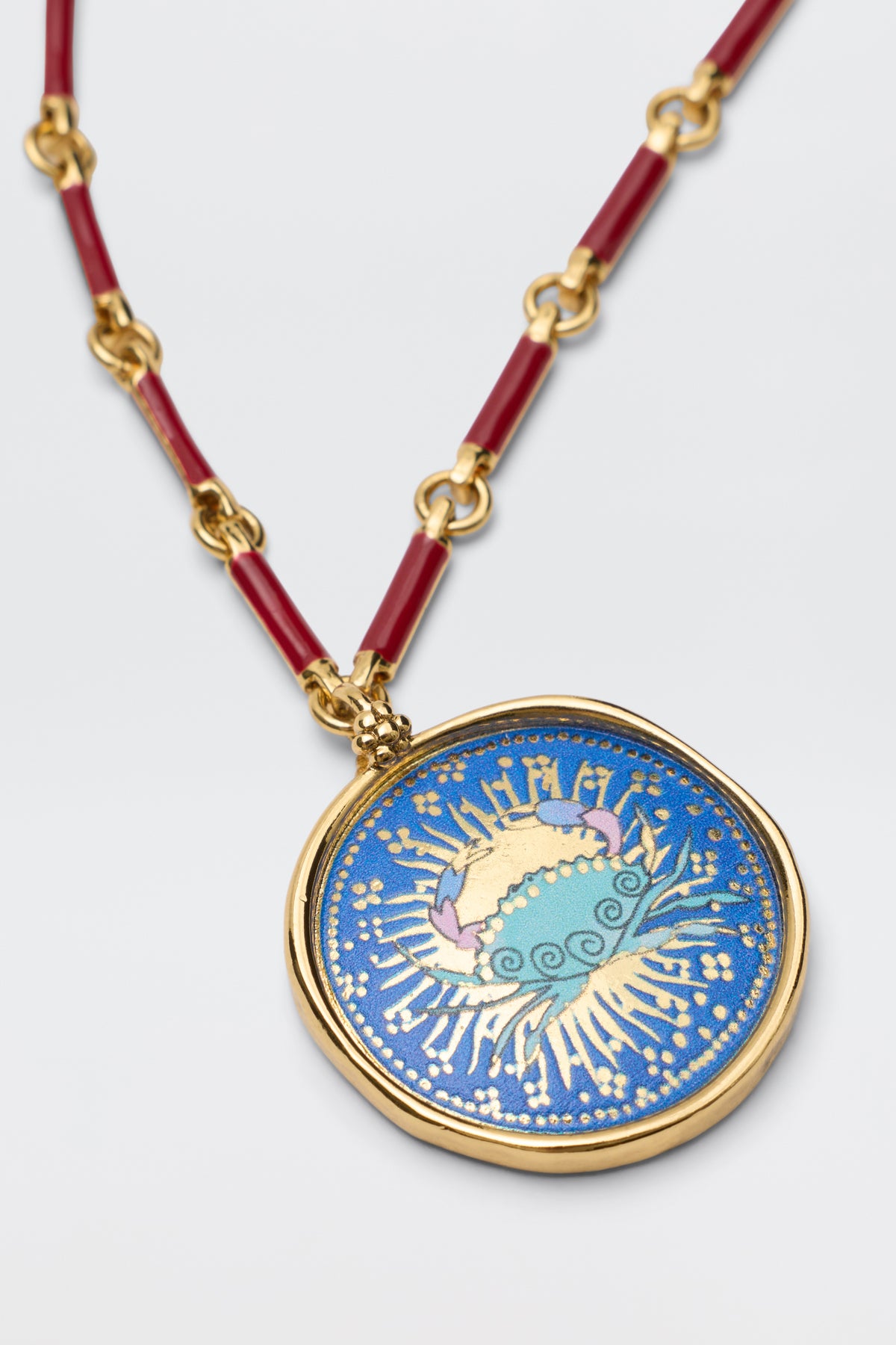 The Ari - Astro Cancer necklace features a round gold disc pendant with blue enamel and abstract yellow, white, and pink patterns. Its chain alternates gold links with red segments, displayed on a light gray background.
