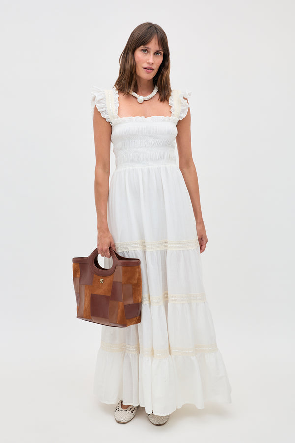 Woman in a long, 70s-inspired ivory nightdress called “Aubrie” with ruffle details holds a brown leather handbag. She wears a chunky white necklace and white shoes, standing against a plain white background.