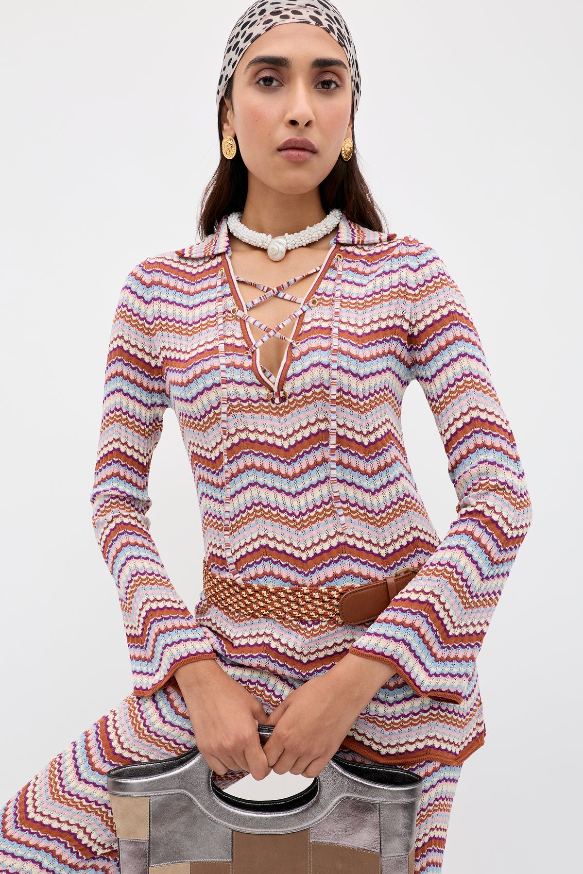 Wearing the Brody - Maria Chevron Multi, a vibrant zigzag-patterned outfit, this woman styles her vintage-inspired look with a lace-up V-neck top, accessorized with a headscarf, shell necklace, and gold earrings. She confidently poses against a plain background while holding a metallic handbag.