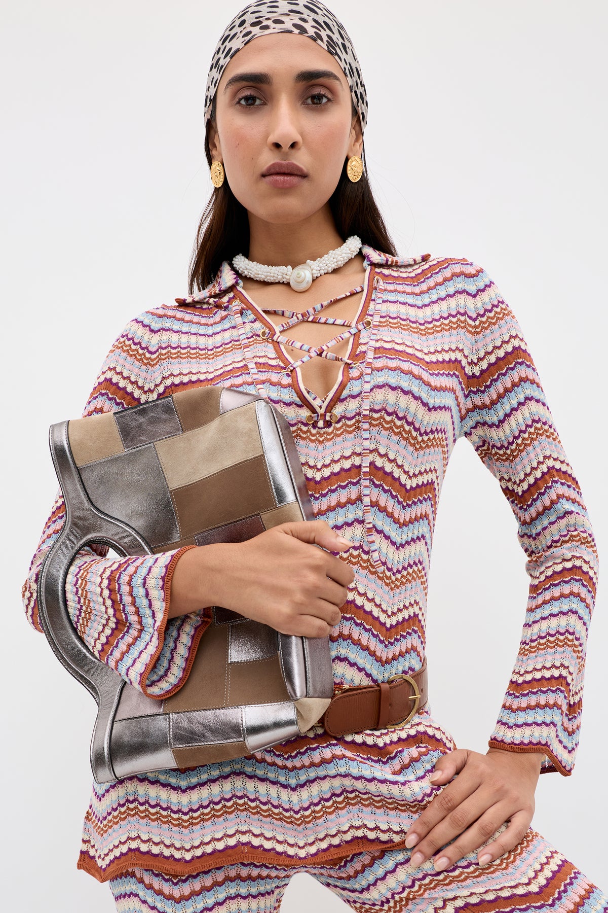 Wearing the Brody - Maria Chevron Multi, a vibrant zigzag retro cotton knit top, this woman completes her look with a patterned headscarf and a patchwork bag. She accessorizes with gold earrings, layered necklaces, and a belt as she poses confidently against a plain background.