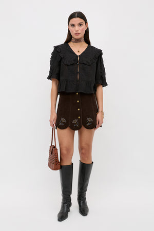 A woman stands against a white background wearing the Caris - Shasta Daisy Embroidery Chocolate mini skirt, paired with a black ruffled blouse, knee-high black boots, a brown woven handbag, and accessorized with jewelry.