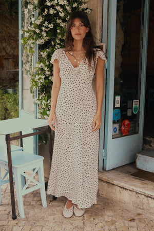 A woman in the Cathy - Layla Dot Ivory dress stands by a light blue chair and table outside a café adorned with climbing flowers and an open blue door behind her.