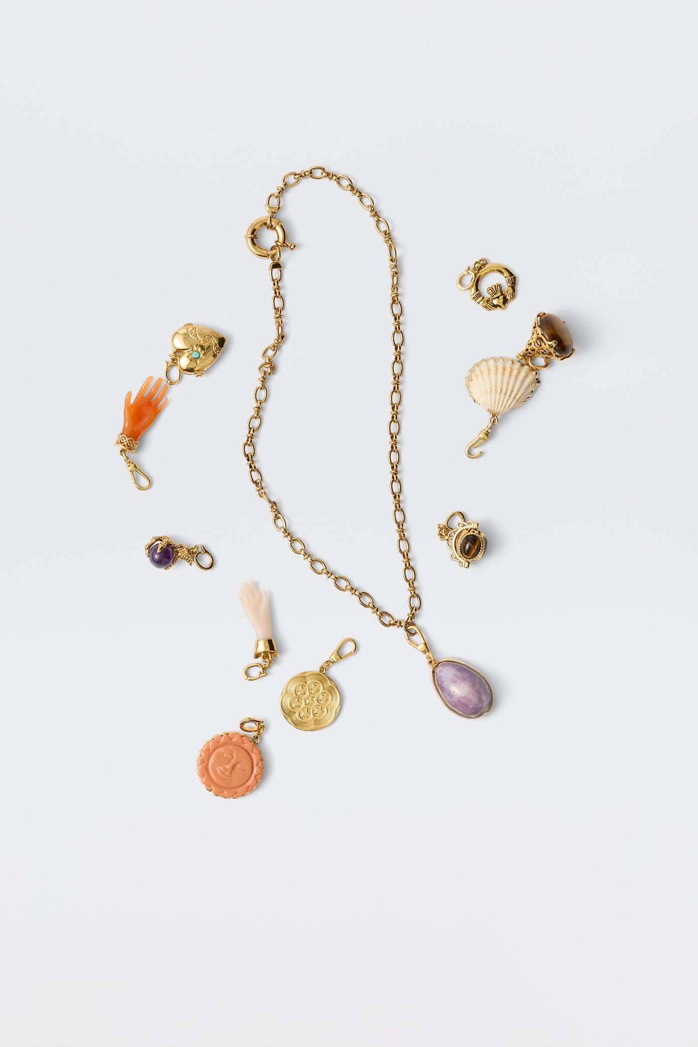 A gold chain Devine necklace with a purple gemstone pendant is displayed on a white background, surrounded by vintage jewellery charms such as the Shell - White charm, an orange hand, coins, amethyst, a tassel, and other decorative pendants.