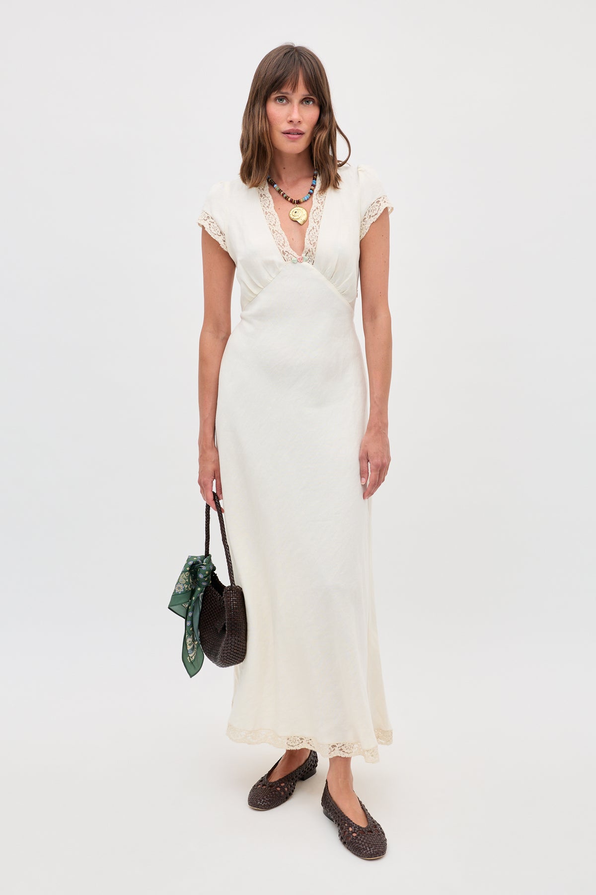 A woman poses against a white background in the Clarice - Cream vintage nightdress with lace trim, accessorized with a dark woven handbag featuring a green scarf, layered necklaces, and dark shoes.