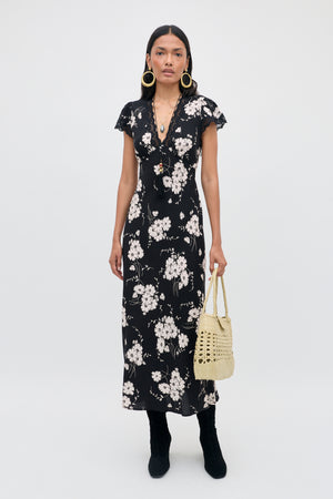 A woman wearing the Clarice - Daisy Field Black dress, paired with black boots, gold hoop earrings, a long necklace, and holding a light-colored woven handbag, stands against a plain white background.
