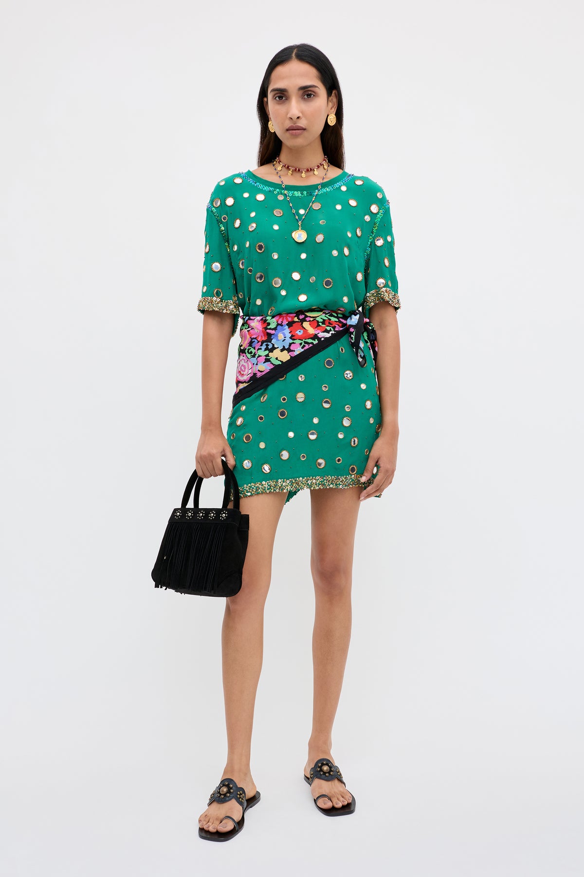 A woman stands against a white background wearing the “Cruz - Lume Embellishment Dark Green” dress, styled with black sandals, gold jewelry, a multicolored floral sash, and holding a black fringed handbag.