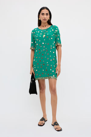 A woman stands against a plain background, wearing the Cruz - Lume Embellishment Dark Green short dress adorned with gold details, paired with black sandals, gold jewelry, and holding a small black handbag.