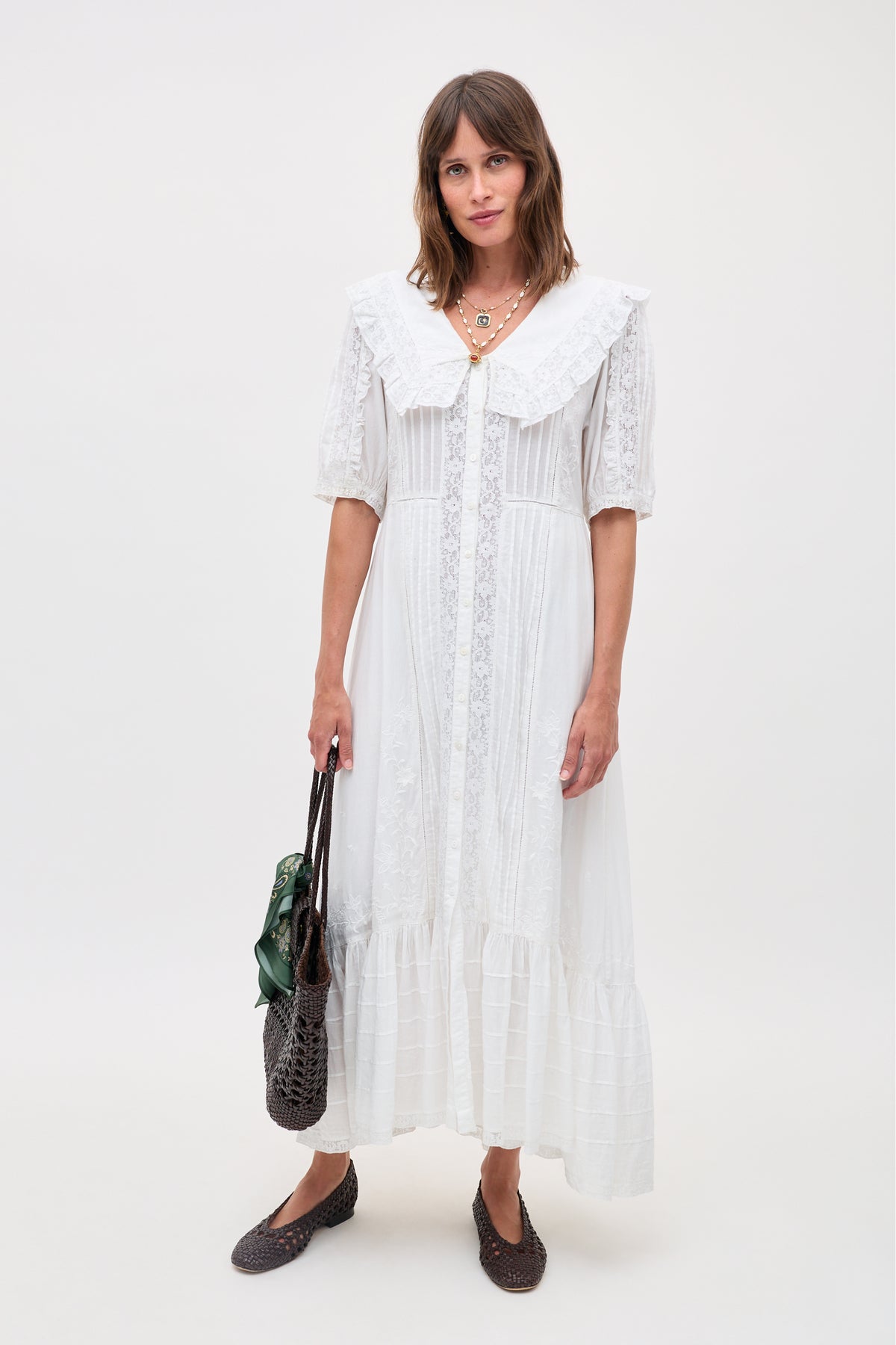 A woman wears the Edina - Pansy Embroidery White long lace blouse dress with puffed sleeves and a large collar, standing against a plain background, holding two bags and wearing brown woven flats.