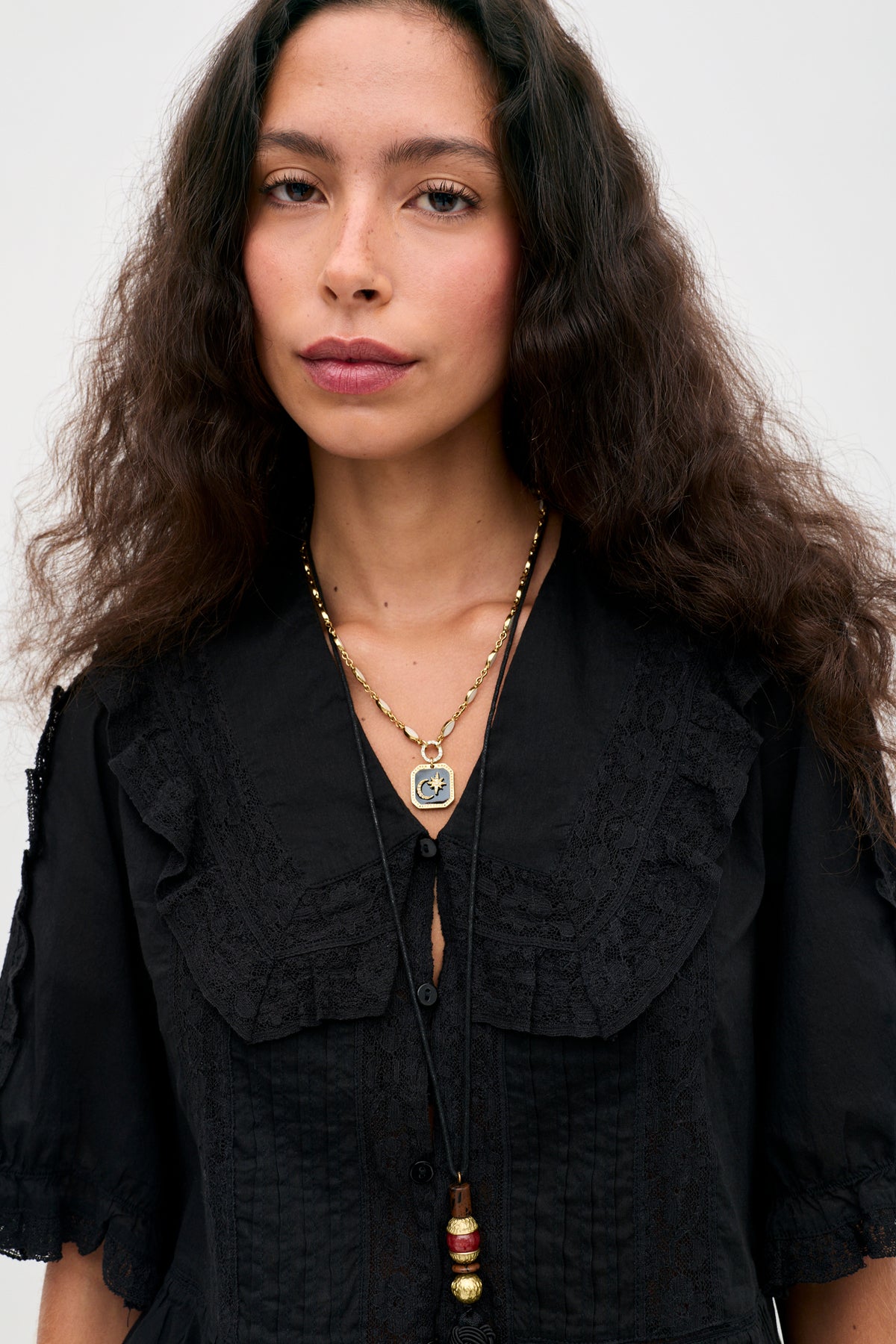 Wearing the “Ellery - Black” vintage-inspired cotton button-down top with ruffled details and a ruffled collar, a woman with long curly hair stands against a plain light background, pairing the look with layered pendant necklaces.