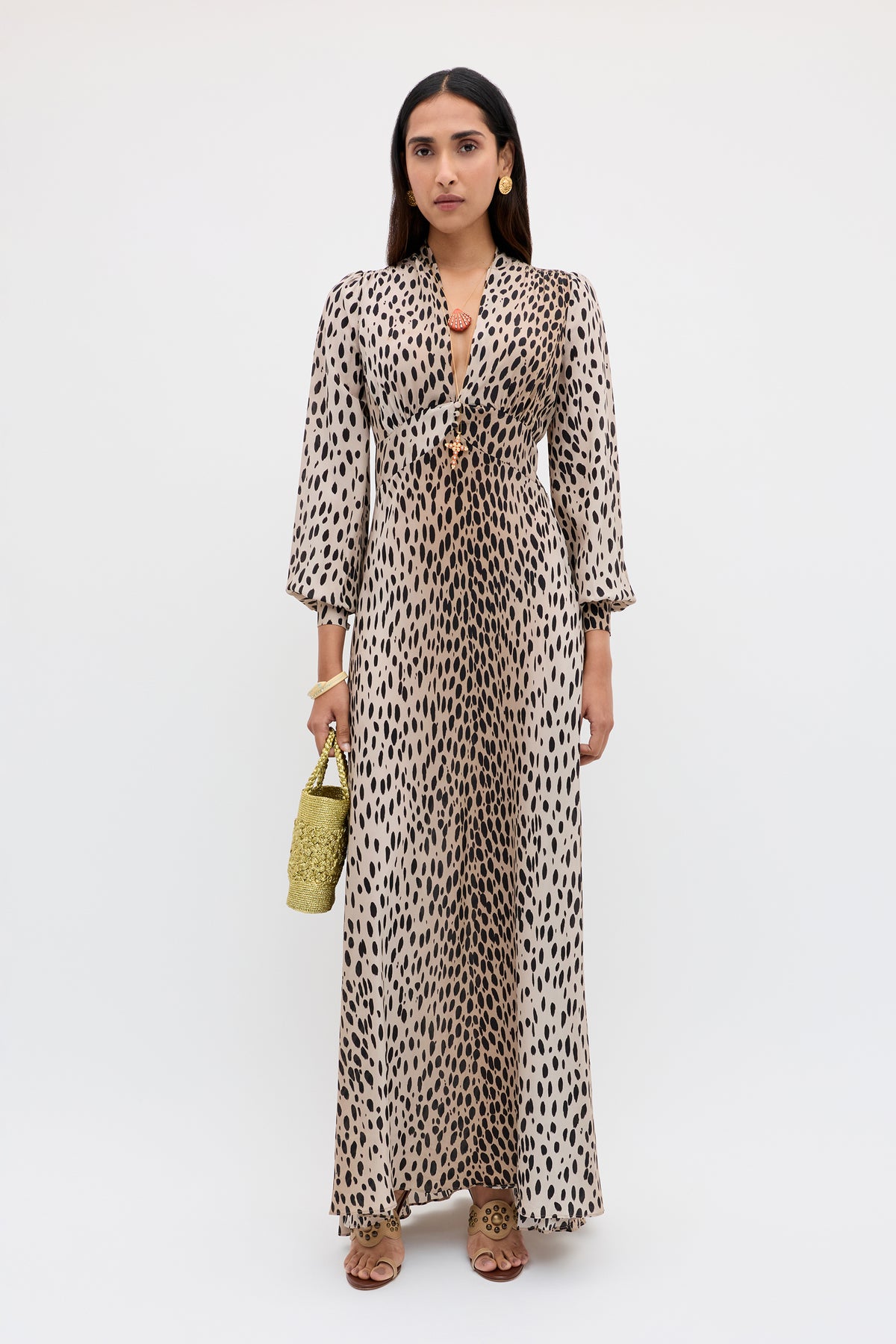 A woman poses in the Emory - Antelope Spot Black silk crepe de chine dress featuring a deep neckline and long sleeves, styled with a green woven handbag, brown heeled sandals, and gold hoop earrings—ideal for special occasions.