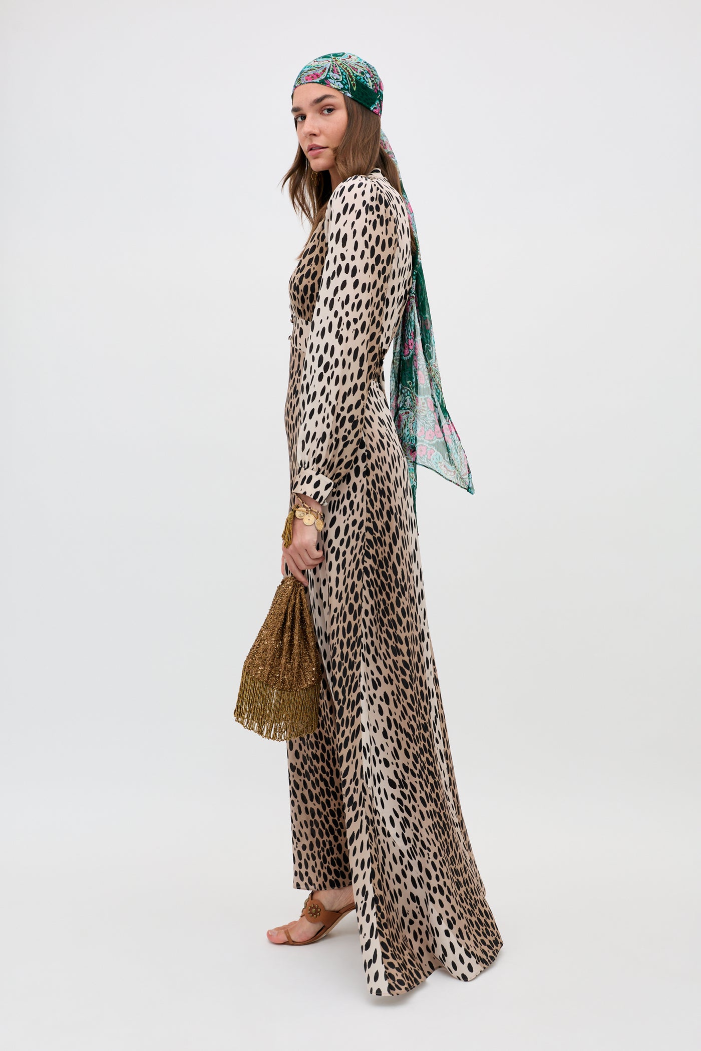 A woman in the Emory - Antelope Spot Black dress and a colorful headscarf stands sideways, holding a textured brown handbag. She wears sandals and gold jewelry, posed against a plain white background.