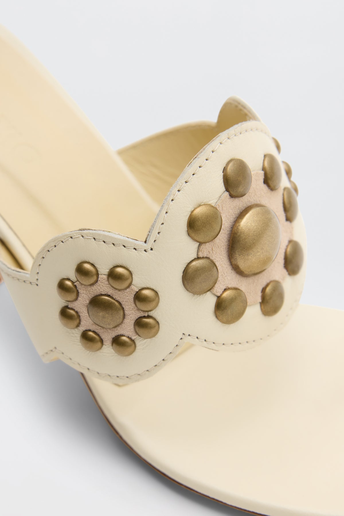 Esmeray - Cream sandal showcases cream-colored straps adorned with gold circular studs and decorative patterns, highlighting a blend of smooth and textured surfaces. The photo emphasizes the intricate detailing of the upper strap.