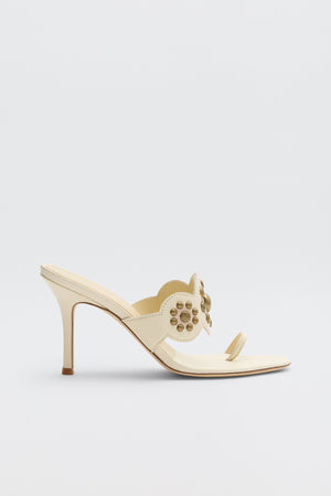 Introducing the Esmeray - Cream: a single cream-colored high heel mule sandal featuring decorative circular cutouts and gold studs on the strap, an open toe design, and a slender stiletto heel, showcased against a plain light background.