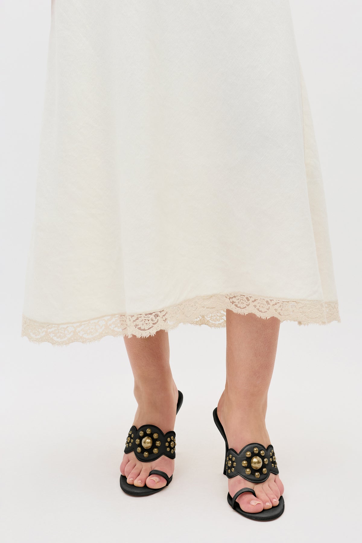 A person wearing a white skirt with lace trim at the hem and the Esmeray - Black sandals, featuring gold studs, standing on a white background.