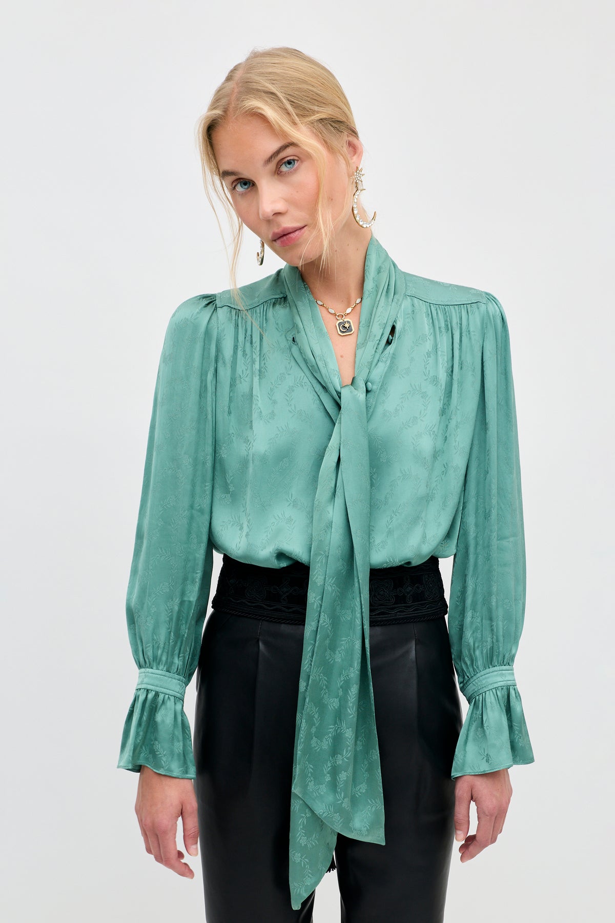 Wearing the Evanora - Floral Trail Jacquard Sage Green blouse, a woman stands against a plain background, looking at the camera. The elegant top features a long necktie and ruffled cuffs, paired with black high-waisted pants for a sophisticated look.
