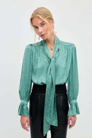 Wearing the Evanora - Floral Trail Jacquard Sage Green blouse, a woman stands against a plain background, looking at the camera. The elegant top features a long necktie and ruffled cuffs, paired with black high-waisted pants for a sophisticated look.