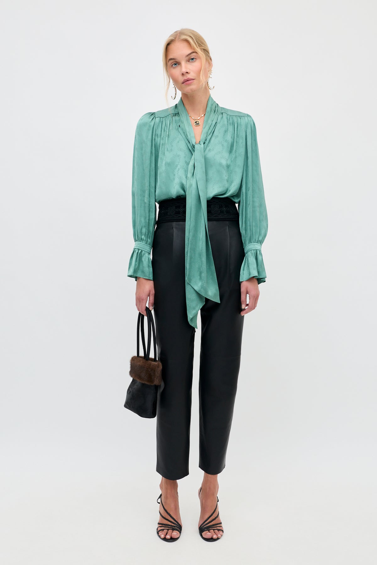 A woman stands against a plain background, wearing the Evanora - Floral Trail Jacquard Sage Green blouse styled with black high-waisted pants, black strappy heels, a small black handbag with fur trim, and statement jewelry.