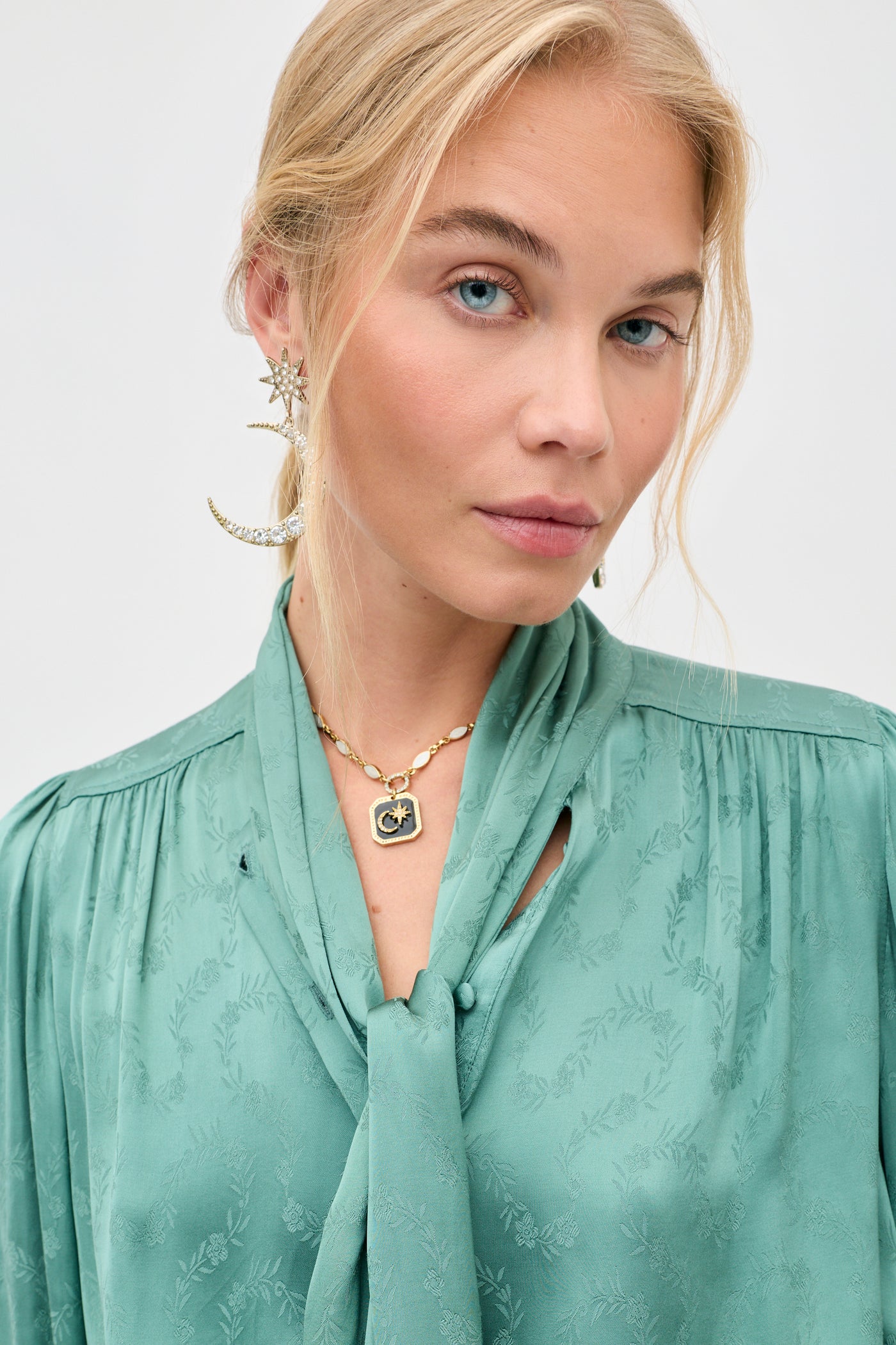 A woman with light blonde hair and blue eyes wears the “Evanora - Floral Trail Jacquard Sage Green” blouse, styled with a necktie, gold crescent moon and star earrings, and a gold necklace featuring a square pendant as she poses against a plain light background.