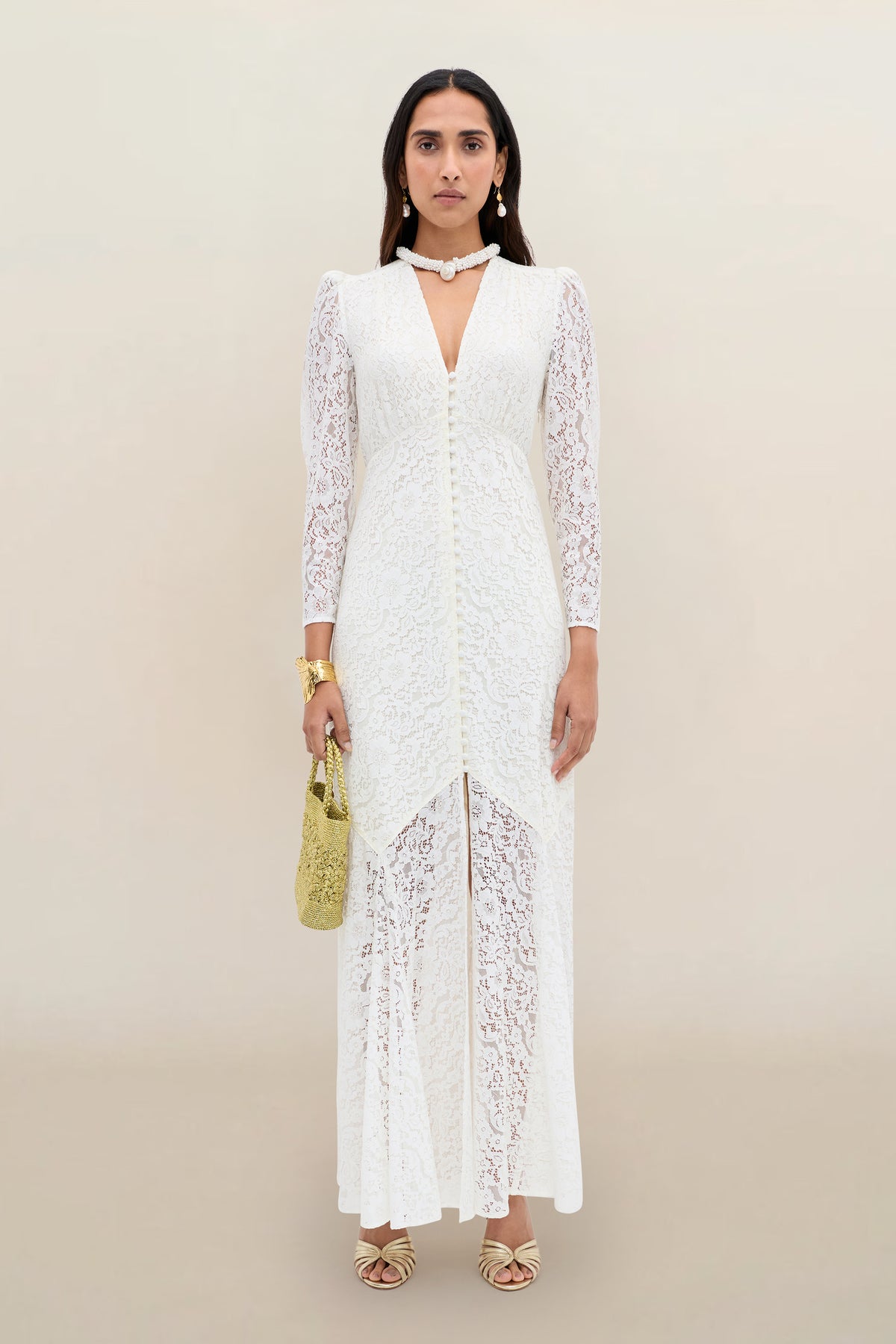 A woman stands against a neutral background wearing the Fleur - Ivory dress, a long white corded lace design with long sleeves and a subtle fit-and-flare silhouette. She completes the look with gold sandals, jewelry, and a gold handbag.