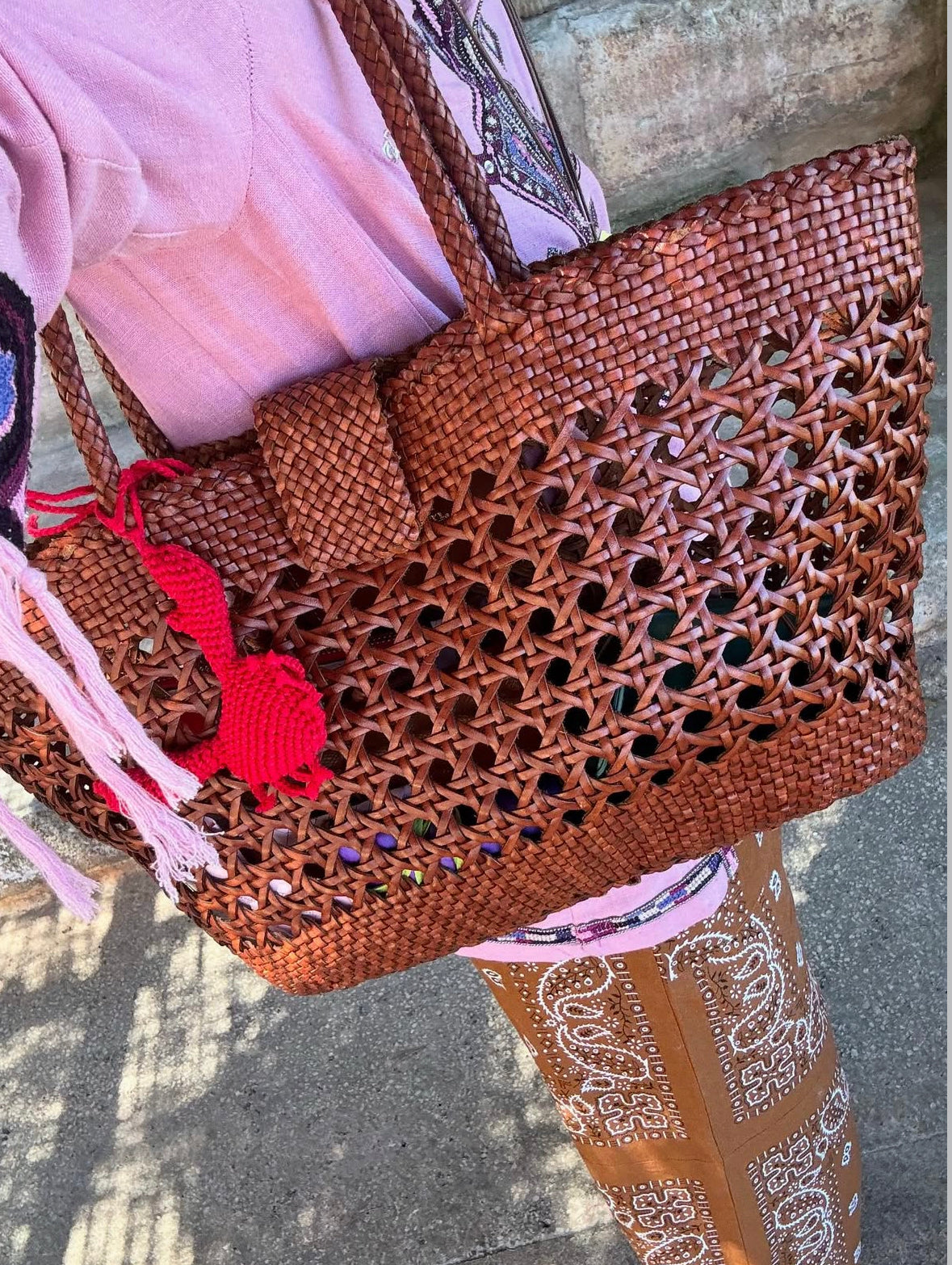 A person in pink and brown patterned clothing carries the Lucine - Tan leather woven bag with a red pom-pom keychain, casting a shadow on the ground.