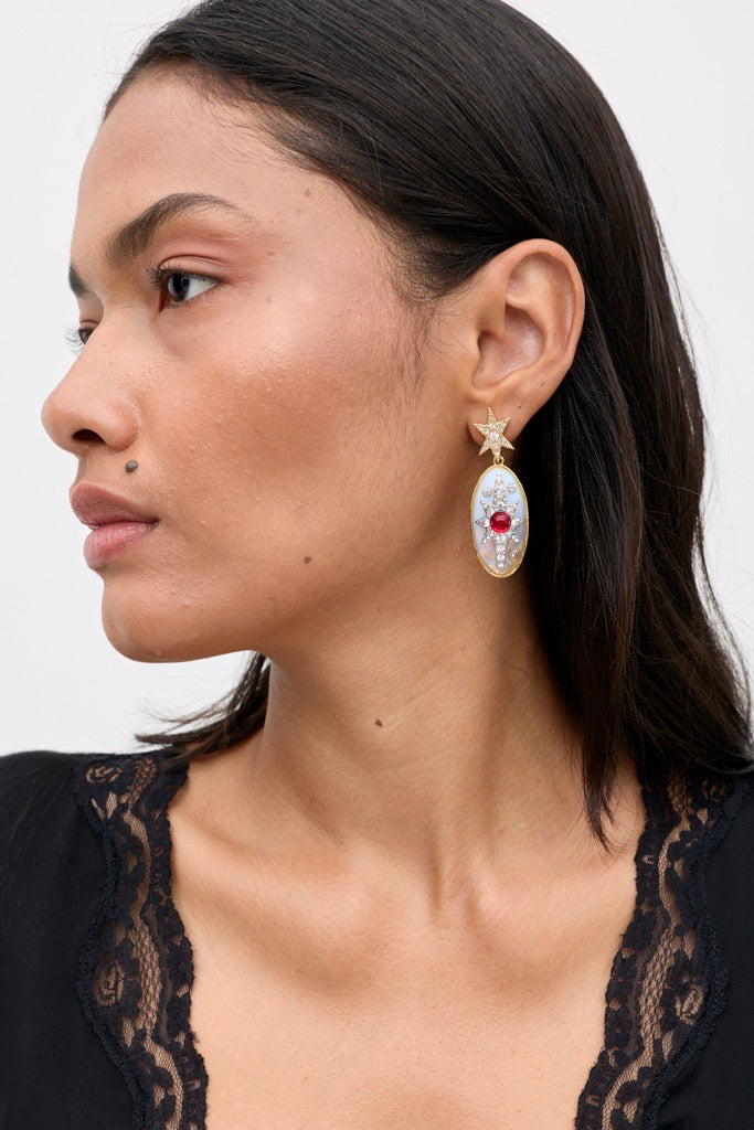 A woman with medium skin tone wears Desiree - Opal, a large, oval-shaped earring featuring a star at the top and gemstone details. She has a beauty mark above her lip and is dressed in a black top with lace trim.