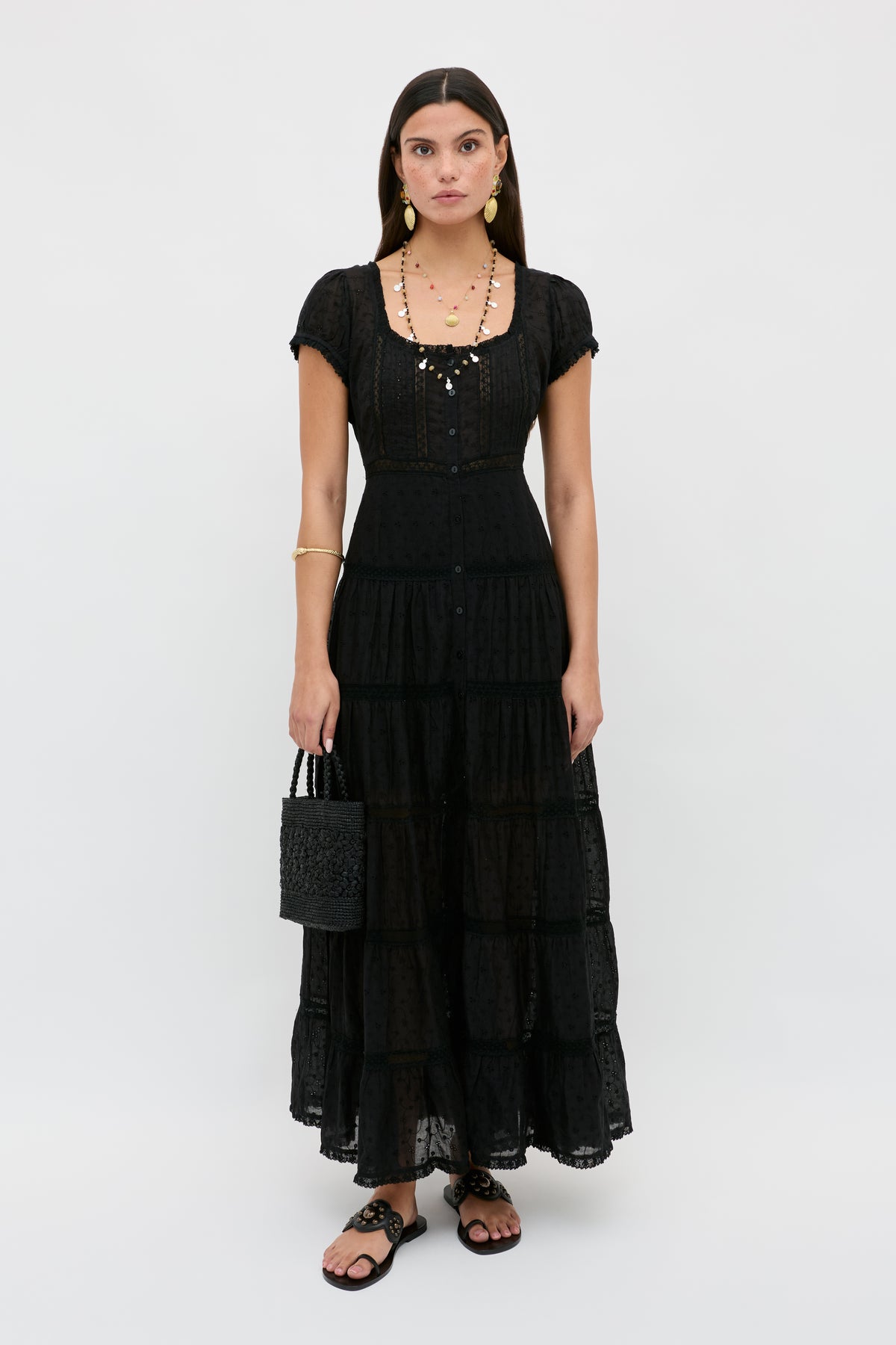 A woman with straight brown hair stands facing forward against a plain white background, wearing the Jaclyn - Black long, tiered black dress with short sleeves. She pairs it with a black woven handbag, black sandals, and gold jewelry.