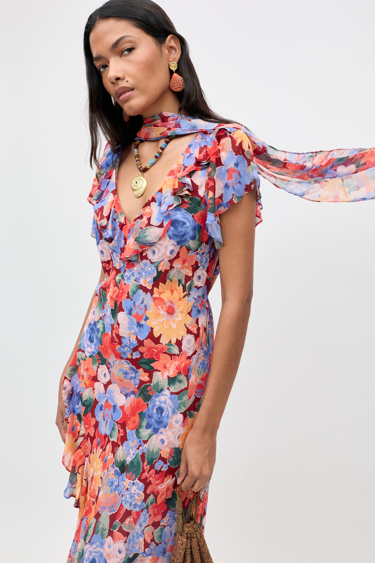 A woman wearing the Julien - Chelsea Floral Red dress, which features vibrant floral patterns and ruffled sleeves, paired with a coordinating scarf. She completes her look with statement earrings, a beaded necklace, and a gold pendant against a plain light background.