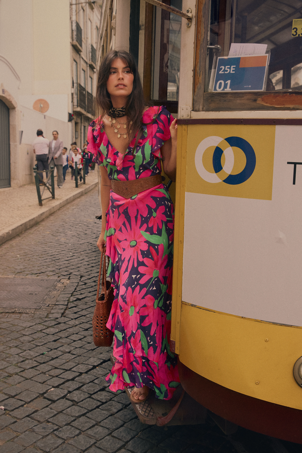 Wearing the Meadow - Danish Floral Pink dress, a woman stands on the steps of a yellow tram along a cobblestone street, holding a brown bag and gazing thoughtfully into the distance.