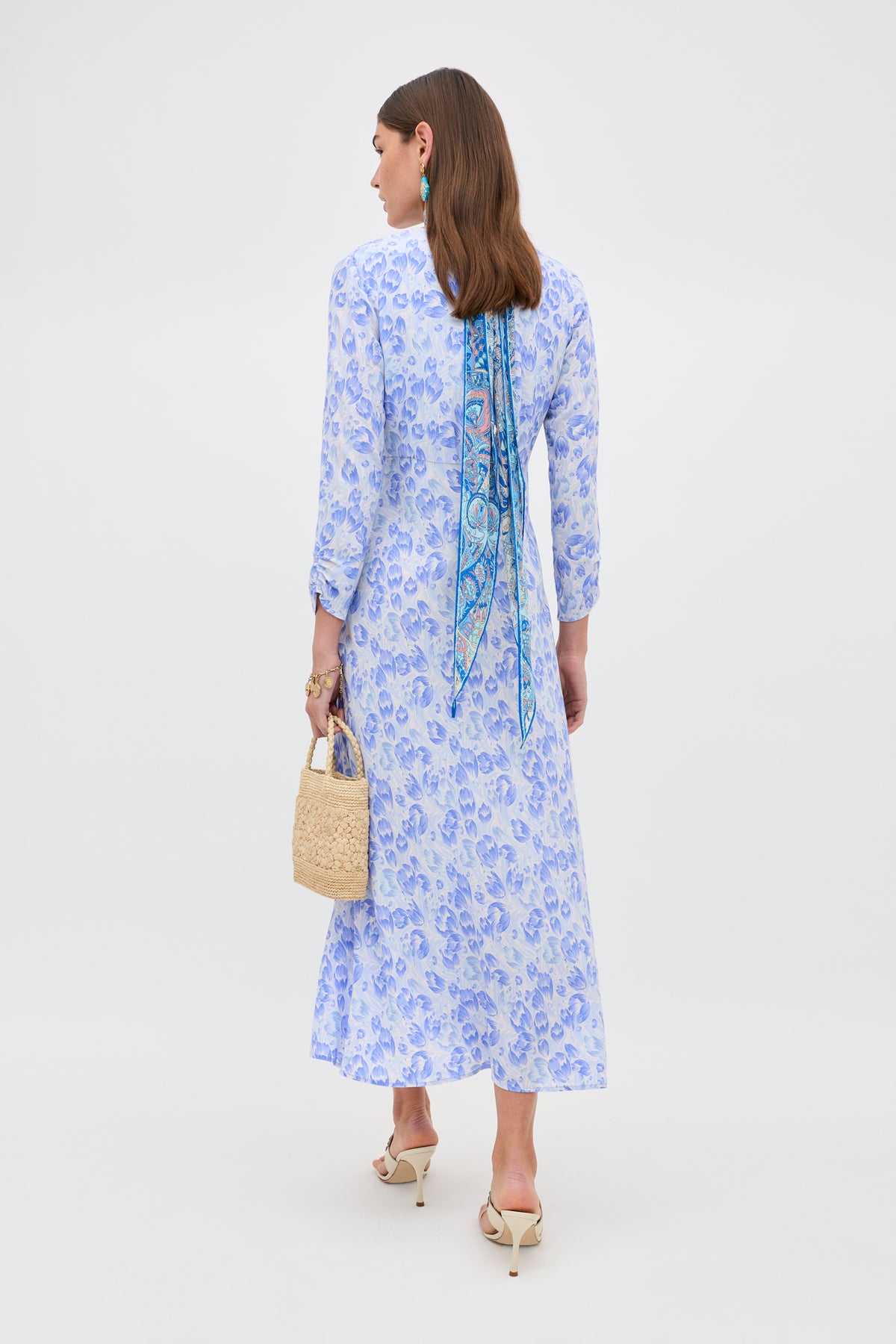 A woman stands facing away, wearing the Katie - Tulip Fields Cornflower Blue dress—ideal for spring and summer. She accessorizes with a woven handbag, patterned hair scarf, white heeled sandals, and statement earrings.