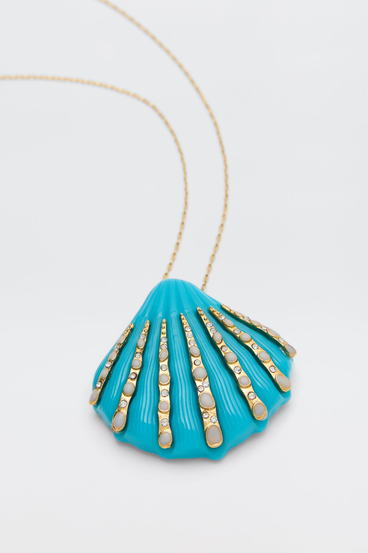 The Leanira - Turquoise is a gold chain necklace with a blue seashell-shaped pendant, accented by gold circular patterned strips, displayed on a plain white background.