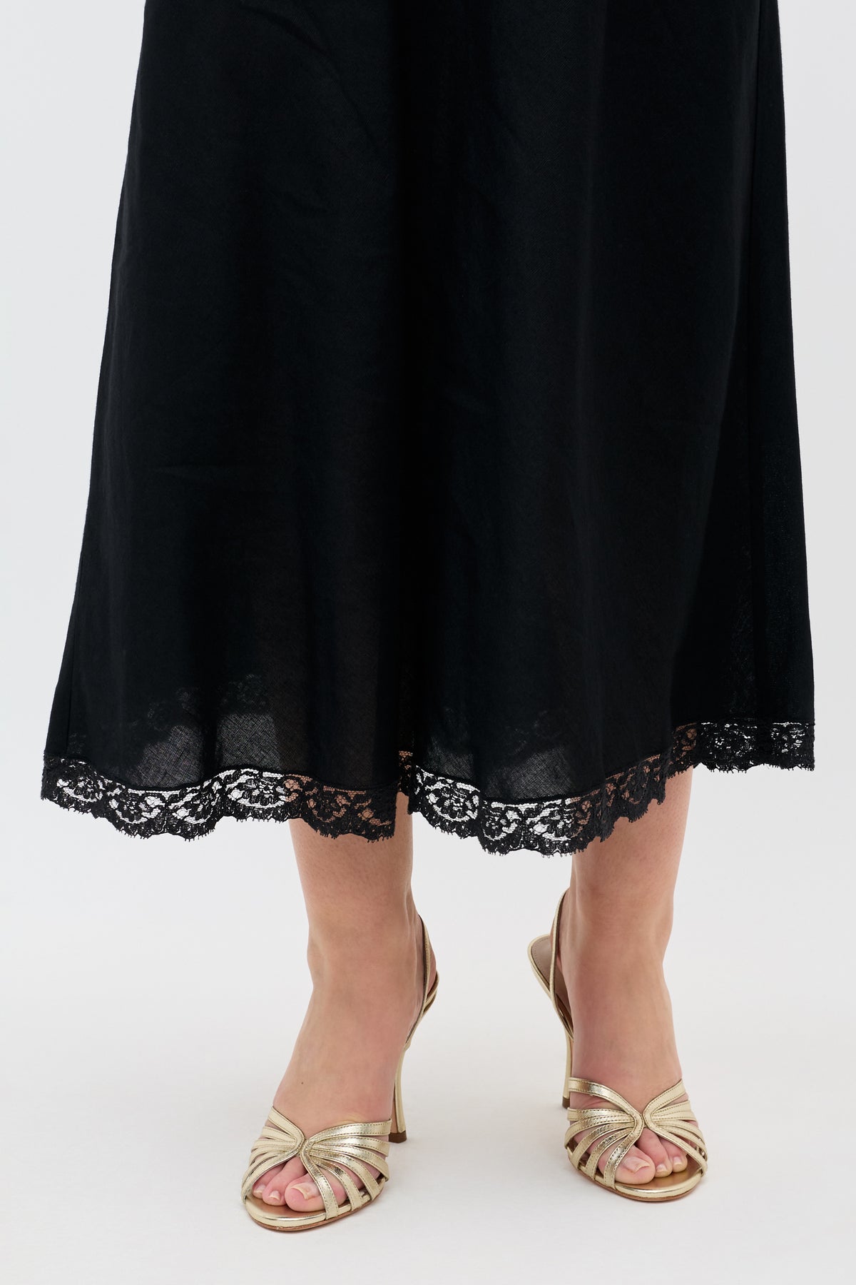 A person wearing a long black skirt with lace trim at the hem and glamorous Mavi - Gold sandals stands on a light-colored surface.