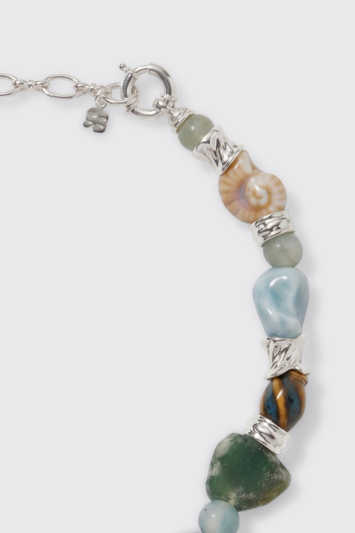 A close-up of the Maxina - Blue necklace, featuring silver links, vibrant polished stones, a shell bead, and a small four-leaf clover charm near the clasp.