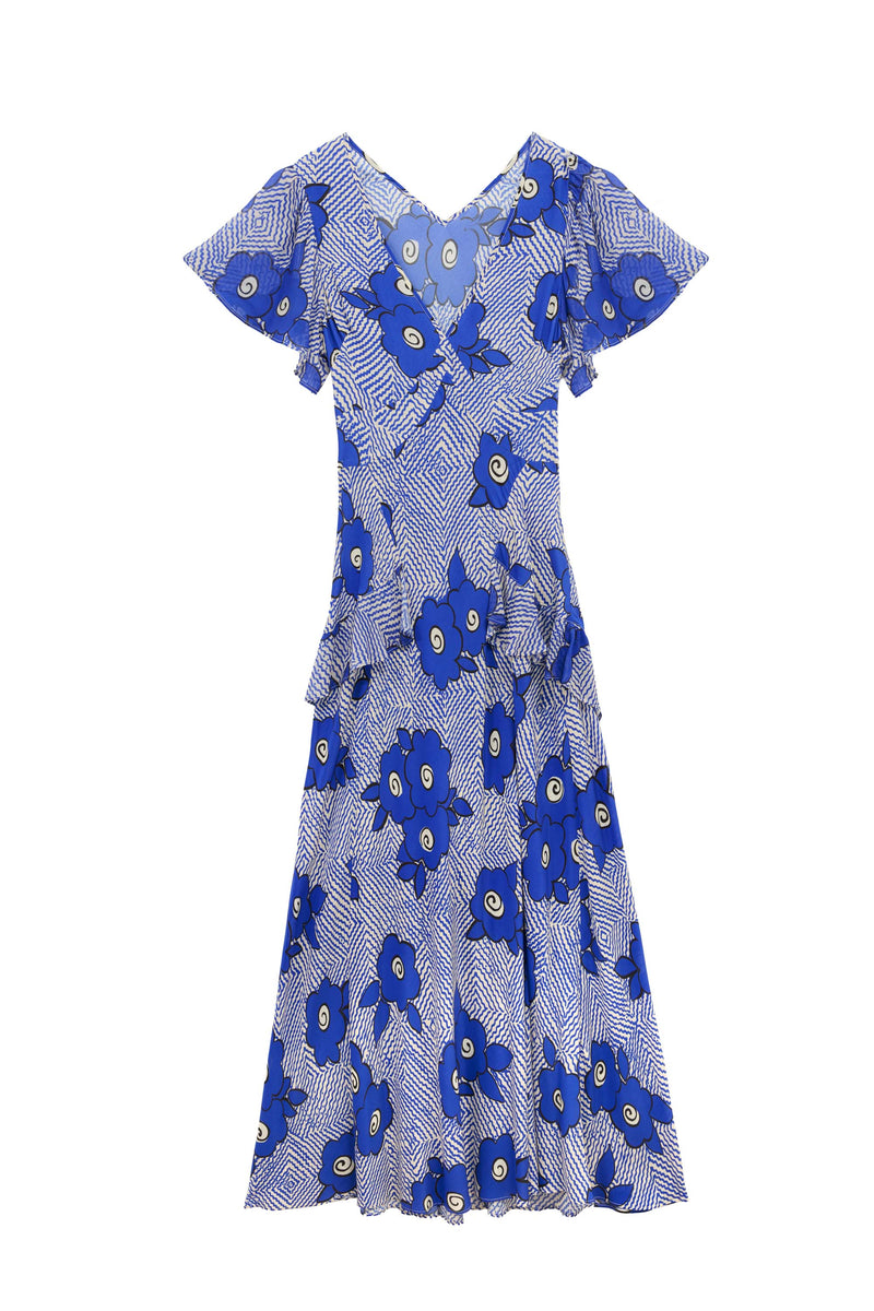 The Evie – Milos Floral Blue dress features a short-sleeve, knee-length silhouette in blue floral print with white geometric lines on a light background, crafted from soft silk crepe de chine and finished with ruffled waist and sleeves.