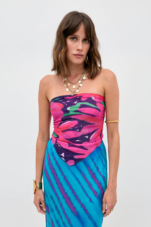 A woman with shoulder-length brown hair wears the Menno - Danish Floral strapless dress, featuring a pink, purple, and blue abstract pattern. She accessorizes with layered gold necklaces, a bangle, and a cuff bracelet against a plain background.