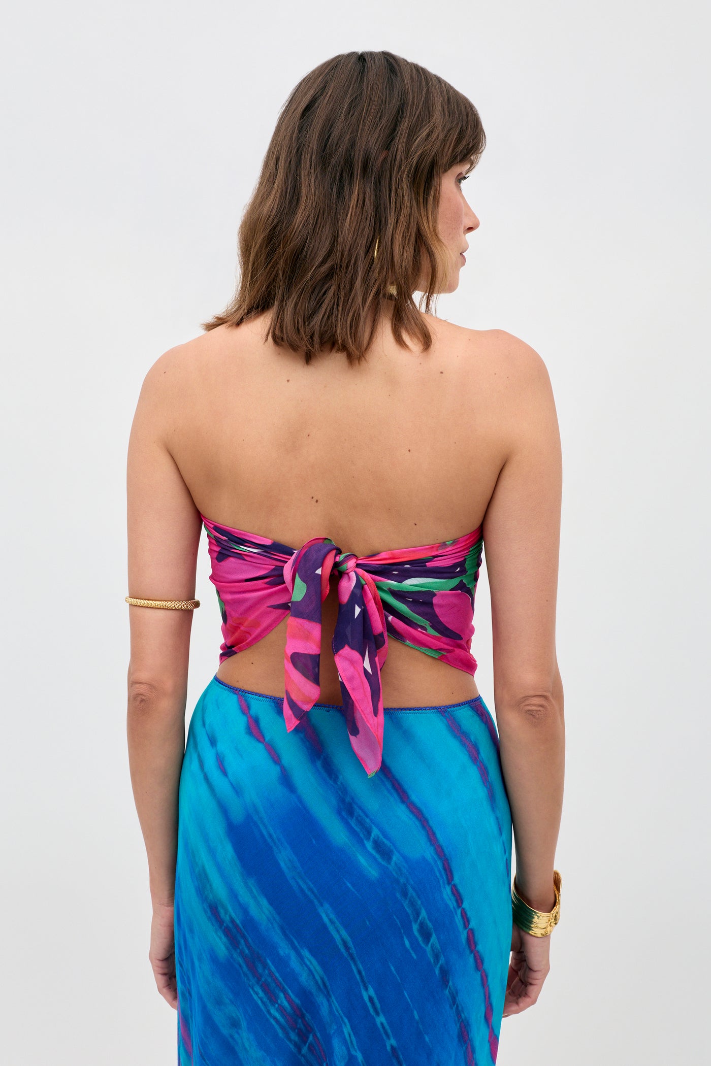A woman with shoulder-length brown hair and gold bracelets on her left arm wears the Menno - Danish Floral strapless top tied at the back with a blue patterned skirt. She faces away against a plain, light background.